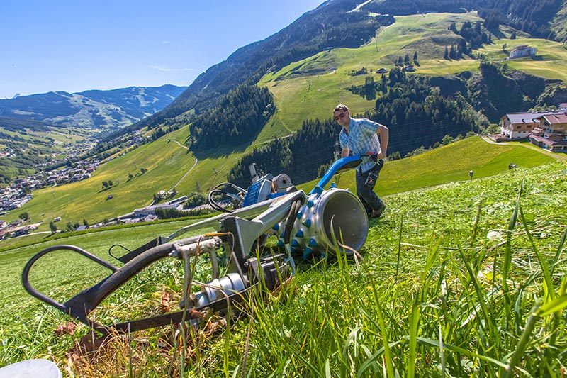 Wolfgang, der Bauer am Perfeldhof, mäht eine steile Wiese mit dem Brielmaier Motormäher. Im Hintergrund erstreckt sich die beeindruckende Bergwelt von Saalbach-Hinterglemm, mit Blick ins Glemmtal. Rechts im Bild ist der Perfeldhof sichtbar, umgeben von der atemberaubenden Naturkulisse.