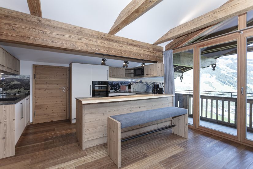 Wohnküche der Ferienwohnung 80 mit Kochinsel, Sitzbank, sichtbaren Holzbalken und großer Fensterfront zum Balkon