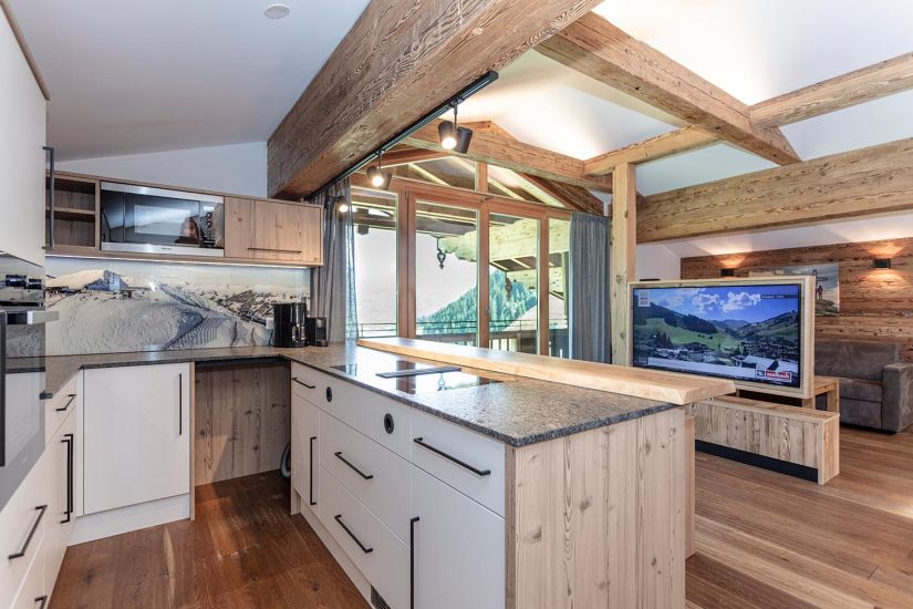 Offene Wohnküche der Ferienwohnung 80 mit Kochinsel, sichtbaren Holzbalken, großem Fenster und Blick ins Tal