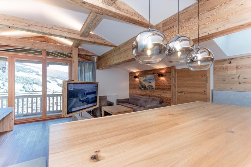 Wohnbereich der Ferienwohnung 80 mit Sofa, Fernseher, sichtbaren Holzbalken, Pendelleuchten und Balkonzugang