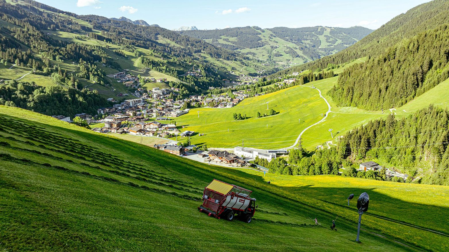 Der Reform Muli fährt die steile Wiese hinunter und nimmt das Silo auf, um es in die Scheune zu transportieren. Im Hintergrund erstreckt sich die sonnige, heiße Landschaft mit einer beeindruckenden Aussicht auf Hinterglemm und das Glemmtal